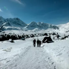 Trzej turyści idą szlakiem przez zaśnieżone Tatry zimą dla średniozaawansowanych, mijając góralskie chatki.