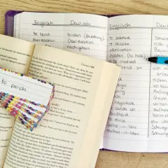 Ein offenes digitales Vokabelheft mit englischen und deutschen Wörtern, daneben ein Buch und Karteikarten zum Lernen.