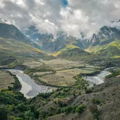 Ein malerischer Fluss schlängelt sich durch ein grünes Tal in Albanien, umgeben von bewaldeten Bergen unter dramatischen Wolken.