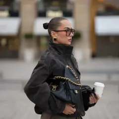 Elegancka kobieta w okularach i ciemnej kurtce z bukli, z torebką Chanel i kawą na wynos.