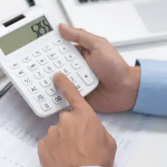 Dłoń naciska klawisze kalkulatora, obliczając wzór na moc prądu. Na stole leżą dokumenty i tablet.