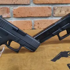 Dwa pistolety Glock, jeden na drugim, na drewnianym blacie z ceglanym tłem. Jeden z nich to pistolet czarnoprochowy Glock.