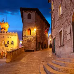 Wieczorna panorama San Marino, z wieżą i kamiennymi budynkami oświetlonymi ciepłym światłem. Niebo mieni się odcieniami różu i błękitu.