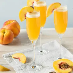 Tre glas med en gyllene drink, garnerade med persikoklyftor. Persikor och en karaff med drinken syns också.