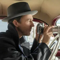 Edward Norton w filmach, w kapeluszu i płaszczu, siedzi za kierownicą samochodu i robi zdjęcie starym aparatem.