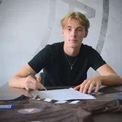 Młody piłkarz Dorian podpisuje kontrakt, obok leży jego brązowa koszulka z logo.