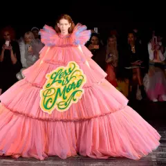 A model wears a voluminous pink tiered gown with a "Less is More" sign, embodying a bold camp aesthetic on the runway.
