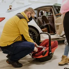 Mężczyzna w żółtej kurtce zapina pasy w czerwonej wkładce do nosidełka Maxi-Cosi, przygotowując ją do podróży samochodem.