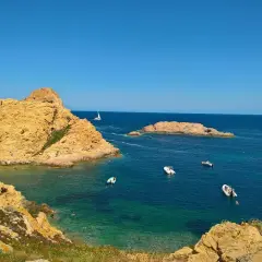 En solig dag vid Ile Rousse. Turkost vatten, klippor och båtar i bukten. En segelbåt syns i fjärran.