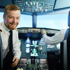 Uśmiechnięty pilot liniowy w kabinie samolotu, obok drugiego pilota obsługującego przyrządy.