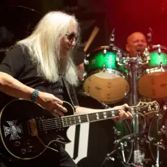 Uriah Heep członkowie na scenie. Gitarzysta z długimi blond włosami gra na czarnej gitarze z symbolem krzyża. W tle perkusista i zielone bębny.