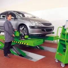 Mechanik przy samochodzie na podnośniku, obok zielona maszyna z monitorem. To profesjonalna tabelą zbieżności kół.
