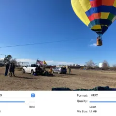 Balon na gorącym powietrzu wznosi się na błękitnym niebie. Na ziemi stoją ludzie, samochody i amerykańska flaga. Obraz porównuje formaty HEIC i JPG.
