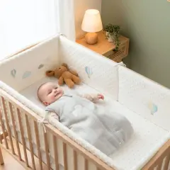 Un bébé souriant dans son lit, entouré d'un tour de lit orné de montgolfières. Un doudou ours est à ses côtés.