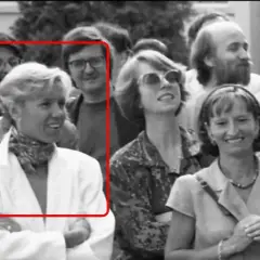 Brigitte Macron młoda, w białej marynarce i z apaszką na szyi, uśmiecha się do aparatu. W tle widać innych ludzi.