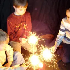 Kinder halten Wunderkerzen. Ein Junge im roten Pullover, ein Mädchen mit Brille und ein kleiner Junge im hellen Pulli genießen den Zauber. Ein schöner Moment, der an die Grundschule erinnert.