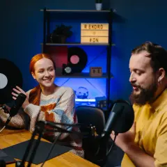 Coppia sorridente registra podcast in inglese interessanti, lei tiene un vinile, lui parla al microfono.