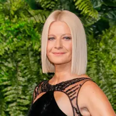 Małgorzata Kożuchowska w czarnej sukni z ozdobnym dekoltem na tle zielonych liści.
