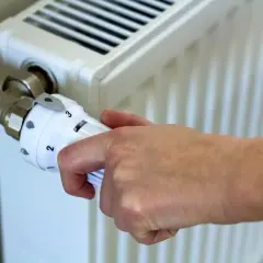 Dłoń reguluje termostat grzejnika, ustawiając temperaturę zgodnie z przepisami dotyczącymi minimalnej temperatury w mieszkaniu.