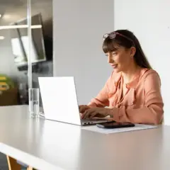 Kobieta pracuje przy laptopie, przygotowując rachunek do umowy o dzieło. Na biurku stoi szklanka wody i telefon.