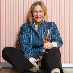 Frida Lund blogg: En glad kvinna med ett glas rosévin sitter på golvet framför en rosa vägg.