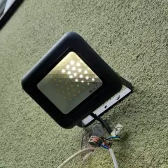 Czarna, nowoczesna lampa naświetlacz LED zamontowana na chropowatej, zielonej ścianie. Widoczne przewody i złączki.