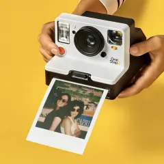 Aparat Polaroid One Step 2 drukuje zdjęcie dwóch kobiet w okularach przeciwsłonecznych na tle kolorowego krajobrazu.
