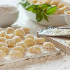 Ñoquis de papa recién hechos, espolvoreados con harina, listos para cocinar. Al fondo, queso rallado y albahaca fresca.
