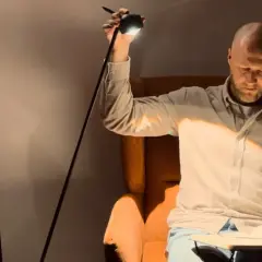 Hombre calvo sentado en sillón leyendo un libro, iluminado por un flexo de pie.