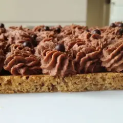 Gâteau rectangulaire avec une base de dacquoise noisette, garni d'une crème au chocolat aérienne et décoré de billes de chocolat.