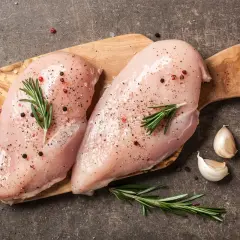 Dwie surowe piersi z kurczaka na desce, przyprawione solą, pieprzem i rozmarynem, obok czosnku.