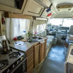 Vue de l'intérieur d'un camping-car américain : cuisine avec cuisinière, évier, et banquette. Drapeaux français et américain visibles.