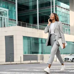 Eleganckie stylizacje do pracy: kobieta w szarym garniturze idzie ulicą, rozmawiając przez telefon i trzymając laptopa.