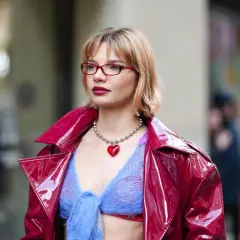 Frau mit roten Brillen und einem stufigen Bob trägt eine glänzende rote Jacke über einem blauen Strickoberteil mit Herz-Halskette.