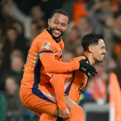 Dwóch piłkarzy Holandii w pomarańczowych strojach świętuje bramkę. Jeden z nich, Memphis Depay, niesie kolegę na plecach, obaj uśmiechnięci.