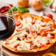 Ein Glas Rotwein neben einer Pizza mit Tomaten und Schinken. Die perfekte Kombination: welcher Wein zu Pizza?