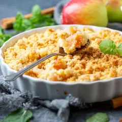Un délicieux crumble pomme doré, garni de feuilles de menthe fraîche. Des pommes rouges et vertes sont visibles en arrière-plan.