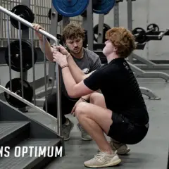 Un coach aide un athlète à améliorer sa **mobilisation articulaire** lors d'un exercice de squat près d'un escalier dans une salle de sport.