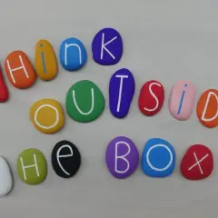 Kolorowe kamienie z literami tworzą napis "THINK OUTSIDE THE BOX". To jak polskie przysłowia po angielsku, zachęcające do nieszablonowego myślenia.