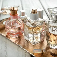 Eleganckie flakoniki perfum, w tym jeden z delikatną różową wstążką, stoją na złotej tacy. W tle błyszcząca biżuteria. Zapach, być może z nutą hexyl cinnamal, unosi się w powietrzu.