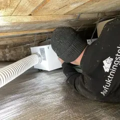 Installering av ventilation i friggebod. En tekniker arbetar med en vit ventilationsenhet under träbjälkar.