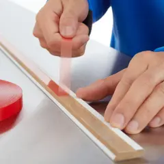 Ręce przykładają cienką taśmę dwustronną do listwy. Obok leży rolka czerwonej taśmy.