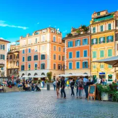 Une place animée à Rome, avec des bâtiments colorés et une fontaine. Un panneau indique "Take Away Cocktail 4.50", un clin d'œil au "saint amour menu".