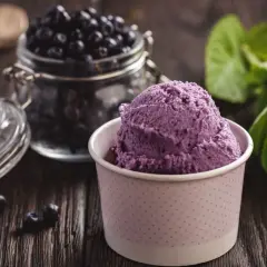 Porcja fioletowego sorbetu borówkowego w papierowym kubeczku, obok słoik z borówkami i liście mięty.