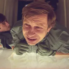 Dwóch mężczyzn w kombinezonach, jeden z przerażoną miną, leży na błyszczącej powierzchni. Scena z filmu "Seksmisja rok produkcji".