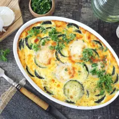 Pyszny obiad z cukinii zapieczony z jajkami i serem. Udekorowany natką pietruszki, obok ser kozi i jajka.
