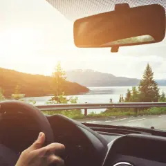 Hände am Lenkrad, Blick durch die Windschutzscheibe auf eine bergige Landschaft mit See. Die Sonne scheint, und man fragt sich, ob man durch das Glas braun wird.