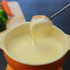 Una forchetta intinge un pezzo di pane in una cremosa fonduta per pasta. Sullo sfondo, verdure fresche.