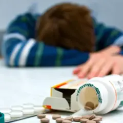 Osoba w bluzie z paskami leży na stole, obok leki, w tym buteleczka z tabletkami, jakby po zażyciu Asentry.