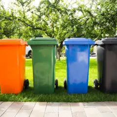 Cztery kolorowe kosze na śmieci (pomarańczowy, zielony, niebieski, czarny) stojące na trawie w ogrodzie.